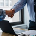 Mod Folder - Crop unrecognizable coworkers in formal wear standing at table with laptop and documents while greeting each other before meeting