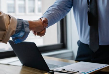 Mod Folder - Crop unrecognizable coworkers in formal wear standing at table with laptop and documents while greeting each other before meeting