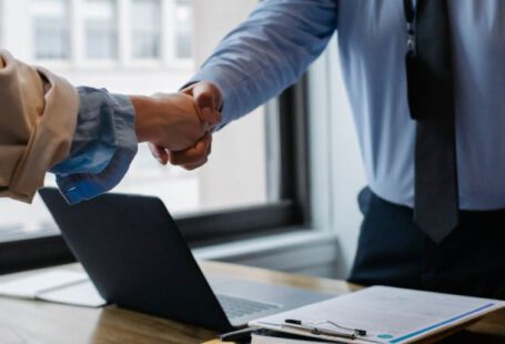 Mod Folder - Crop unrecognizable coworkers in formal wear standing at table with laptop and documents while greeting each other before meeting