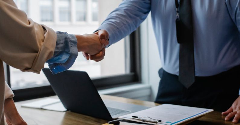 Mod Folder - Crop unrecognizable coworkers in formal wear standing at table with laptop and documents while greeting each other before meeting