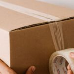 Service Pack - Crop anonymous male using tape to seal packed cardboard box against white wall while moving into new place