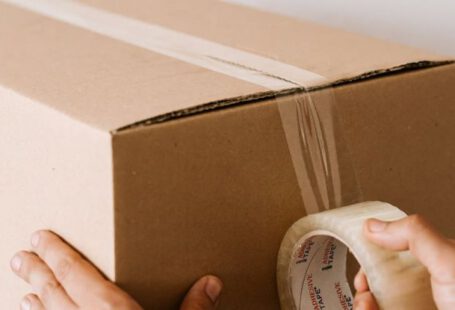 Service Pack - Crop anonymous male using tape to seal packed cardboard box against white wall while moving into new place