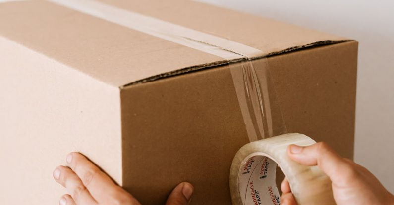 Service Pack - Crop anonymous male using tape to seal packed cardboard box against white wall while moving into new place